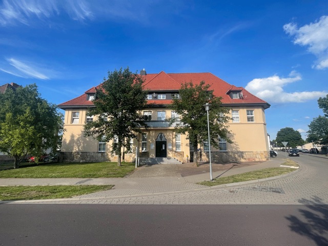 Das Landambulatorium von vorn: gelbes Gebäude mit rotem Dach, Eingangstreppe und Bäumen, rechts die Straße